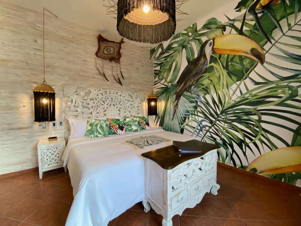 Interior view of a bedroom at Centro Tulum 6 Room Hotel in Tulum, featuring a large mural of tropical leaves and toucans, a carved white wooden bed frame, and woven hanging lamps.