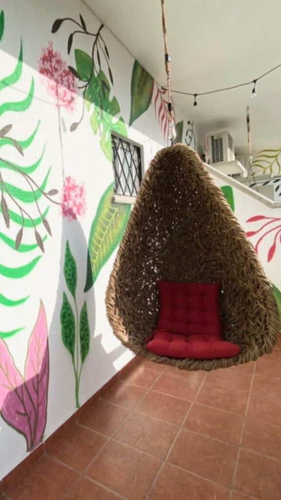 A hanging wicker chair with a red cushion is indoors at Centro Tulum in Tulum, featuring a colorful mural of green and pink leaves and flowers on the wall.