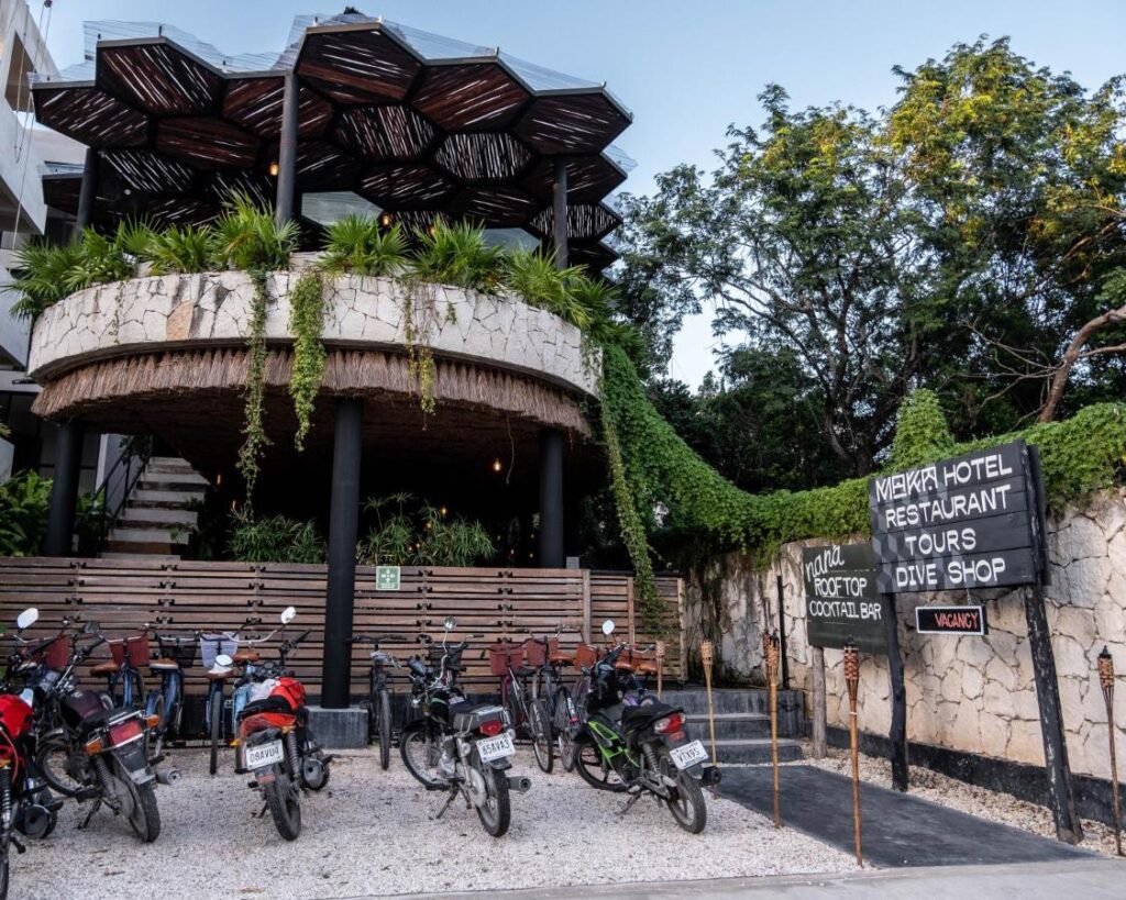 The Maka Front building at Centro Tulum 20 Room Hotel in Tulum features a distinctive hexagonal rooftop structure surrounded by lush greenery, with motorcycles parked in front and signs for the hotel’s amenities.