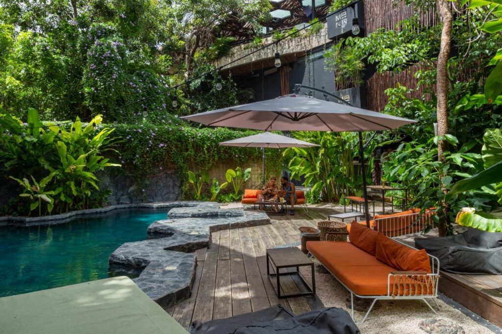 A relaxing outdoor scene at Maka Pool in Centro Tulum 20 Room Hotel, Tulum, featuring a swimming pool surrounded by lush greenery, outdoor seating, and shaded umbrellas.