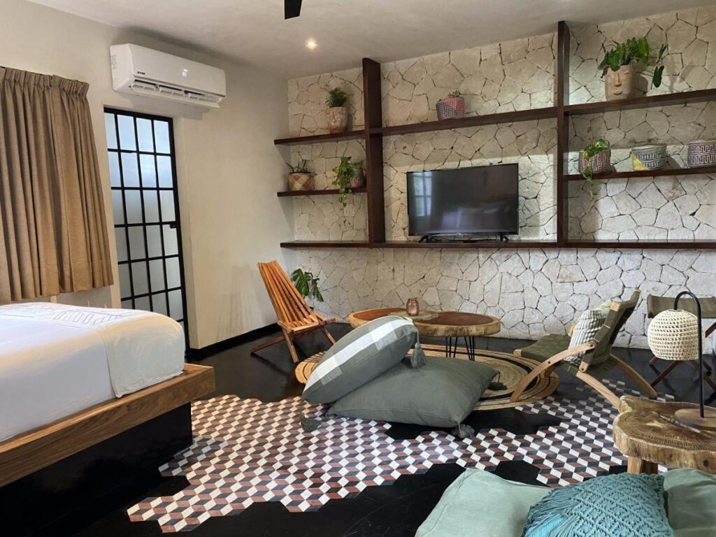 Interior view of the Maka Room at Centro Tulum 20 Room Hotel in Tulum, featuring a stone accent wall, wooden shelves with plants and decor, and a cozy seating area.