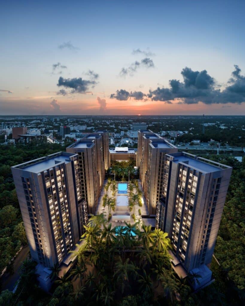 Aerial view of Manglar Cumbres L in Cancun at sunset, showing modern high-rise buildings, a swimming pool, and lush landscaping amidst a cityscape and distant horizon.