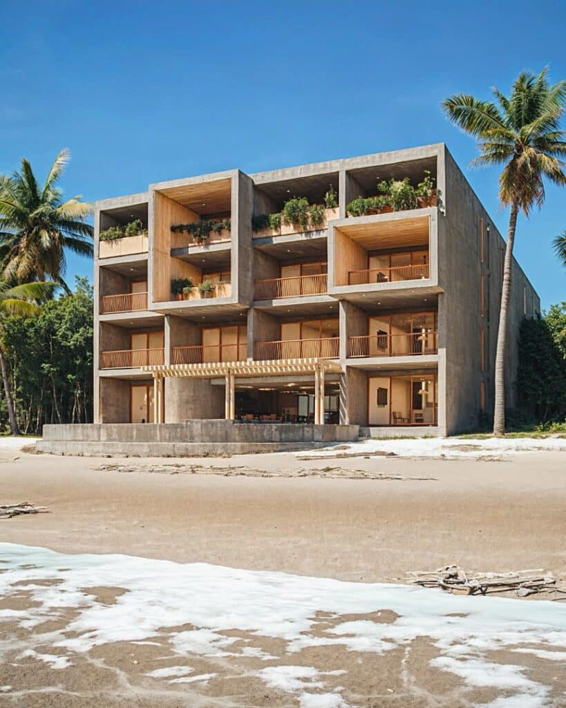 A modern, concrete beachfront building at Maranta 203 Beachfront in Tulum features multiple levels with balconies and lush greenery, situated directly on the sandy shoreline.