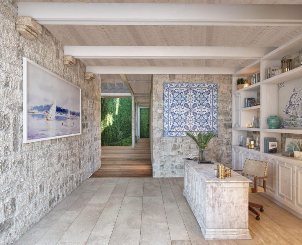 Interior view of Puerta Azul Tulum 101 in Aldea Zama, Tulum, showing a textured stone wall, wooden ceiling beams, a bookshelf with decor, and a corridor leading outside.
