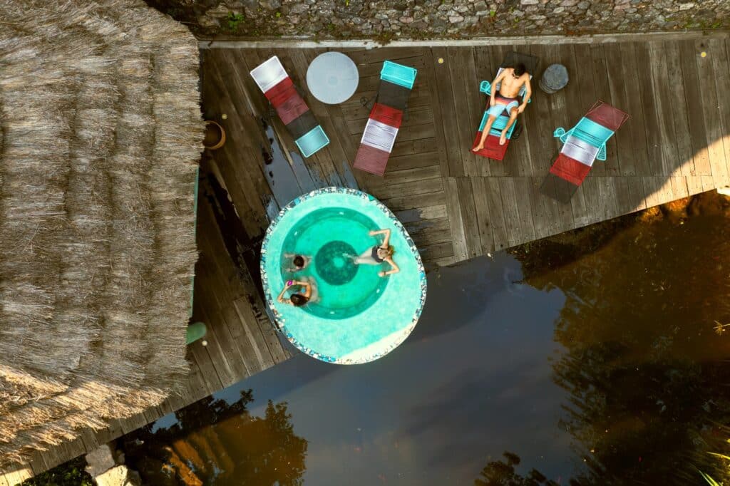 A view from above shows two women relaxing in a hot tub on a wooden deck surrounded by water, with colorful lounge chairs and a thatched roof nearby in Cerro Colorado, Loma De Rodriguez.