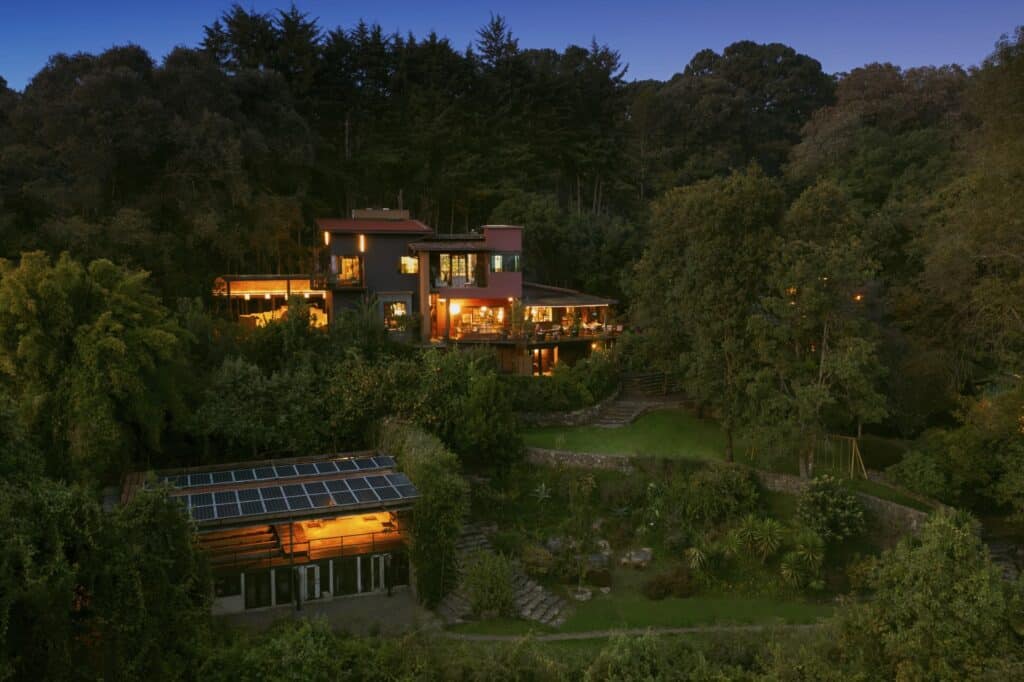 A modern multi-level house with large windows and outdoor terraces is surrounded by lush greenery in Cerro Colorado, Loma De Rodriguez, illuminated at dusk.