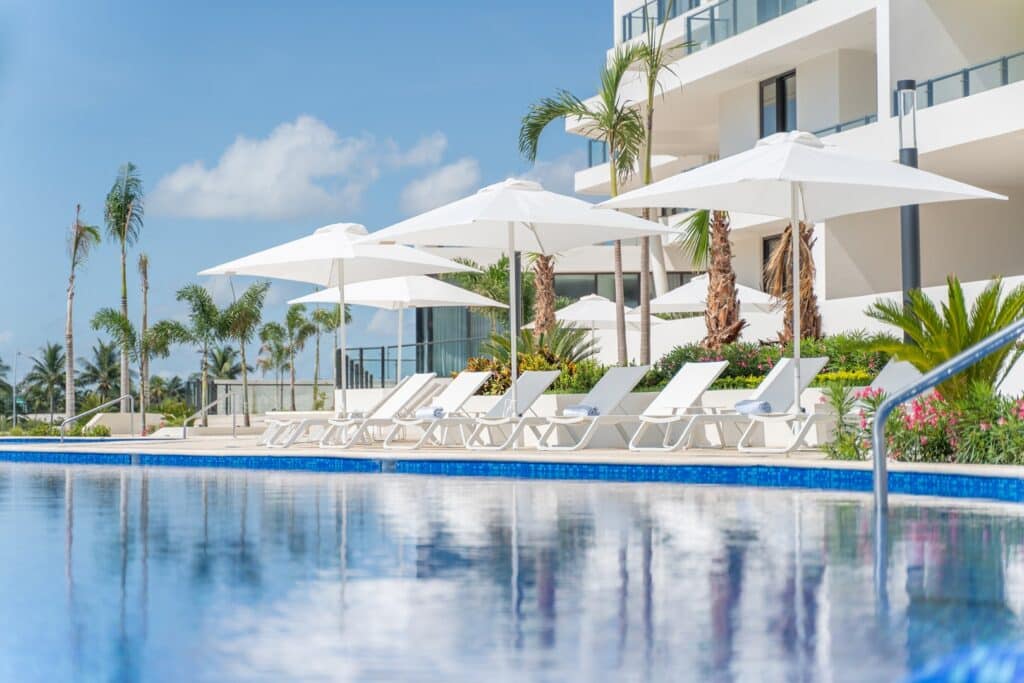 A modern swimming pool area at Blume 1C in Puerto Cancun, with white lounge chairs, umbrellas, and lush tropical plants beside a sleek residential building.