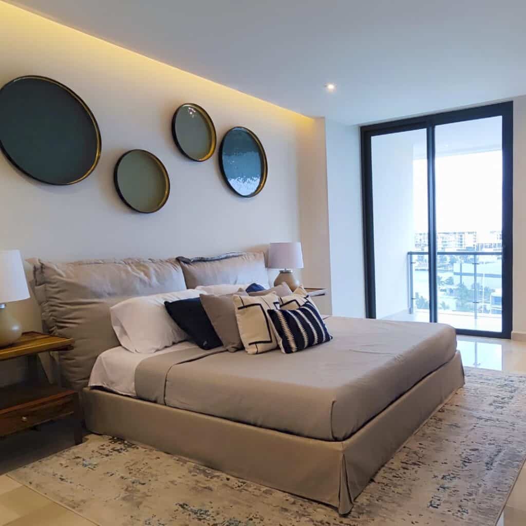 A modern bedroom in Blume 1C development, Cancun, featuring a large bed with neutral-colored bedding, decorative pillows, and a wall decor of circular mirrors, with a glass door leading to a balcony.