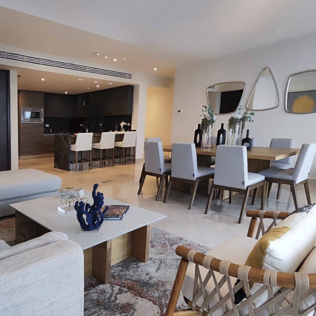 Interior shot of a modern living and dining area in Blume 1C, Puerto Cancun, featuring neutral-toned furniture, decorative mirrors on the wall, and an open kitchen.