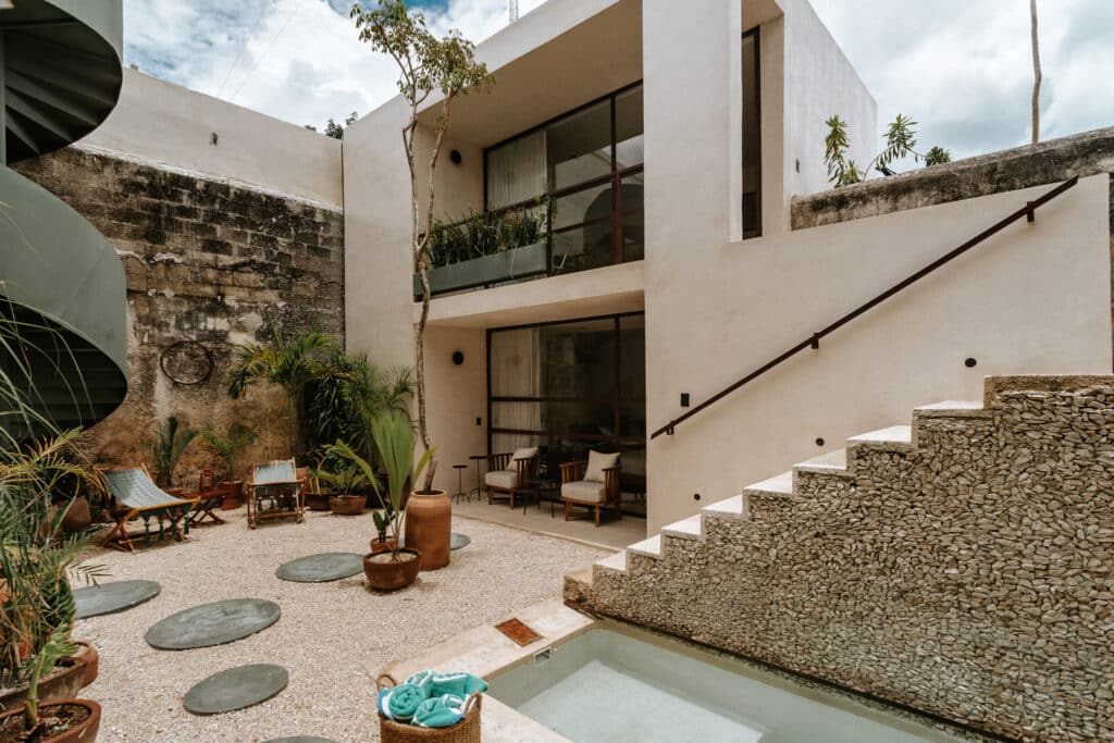 A modern two-story home in Axiotic Villa, Centro Merida, with an outdoor patio featuring potted plants, seating area, gravel ground, and a small pool under a partly cloudy sky.