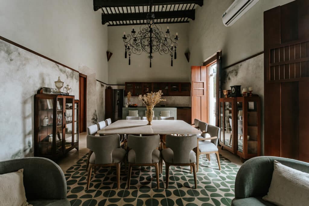 A dining area in Axiotic Villa in Centro Merida features a large table with a floral centerpiece, vintage furniture, green patterned tile flooring, dark wood cabinetry, and a wrought iron chandelier.