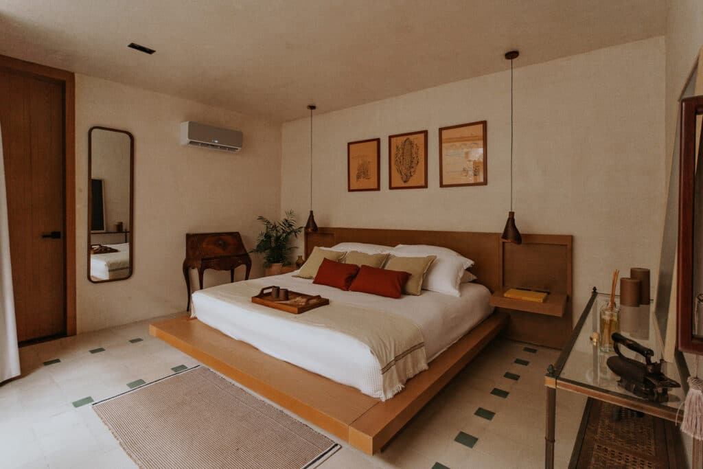 A bedroom at Axiotic Villa in Centro Merida features a wooden bed with neutral-toned pillows, framed artwork above, a vintage writing desk, and warm, minimalist decor.