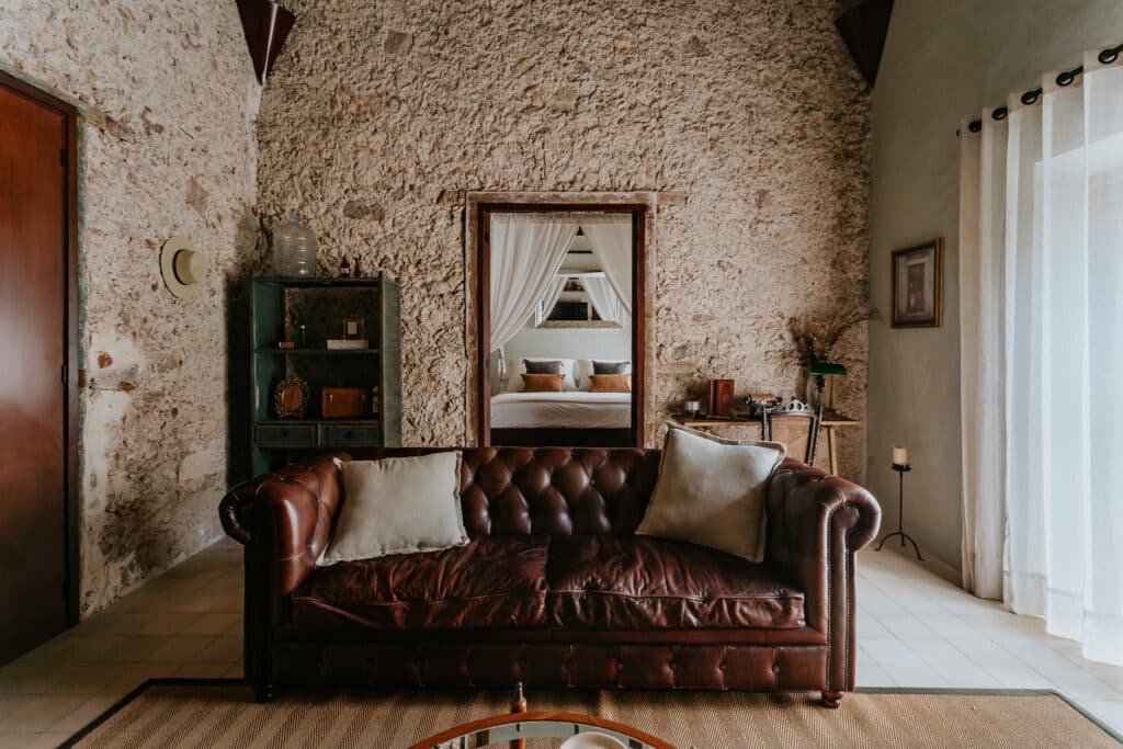 Living room of Axiotic Villa in Merida’s Centro Merida with a leather sofa, stone walls, vintage furniture, and sheer curtains overlooking a bedroom through a doorway.
