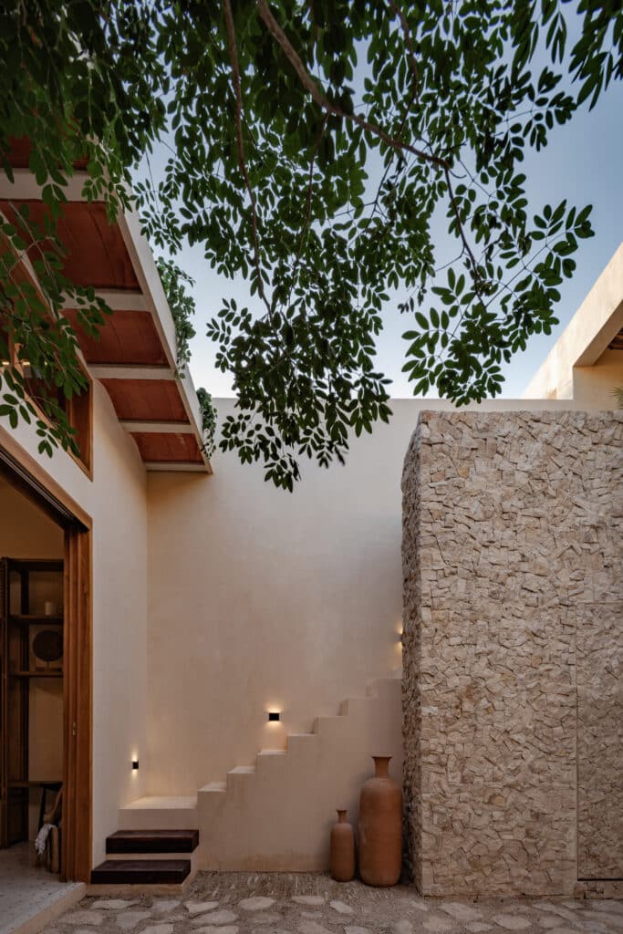 A modern courtyard at Casa Caracol Merida Villa 19 in Caracol 525, Centro Merida, featuring a stone wall, wooden accents, and lush tree branches overhead.
