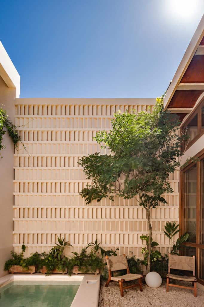 A view of Casa Caracol Merida Villa 20 in Centro Merida, featuring a modern outdoor area with a small pool, wooden chairs, and a decorative beige wall under a bright blue sky.