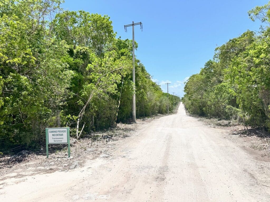 A dirt road with utility poles on each side runs through lush green vegetation in Land, Puerto Aventuras, Xpu Ha, with a sign indicating a private, no entry pathway.