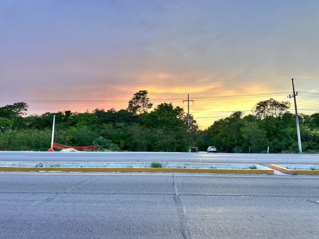 A plot of land in Land development near Xpu Ha, Puerto Aventuras, with a sunset sky and trees in the background, visible from the street with a guardrail and parked cars.