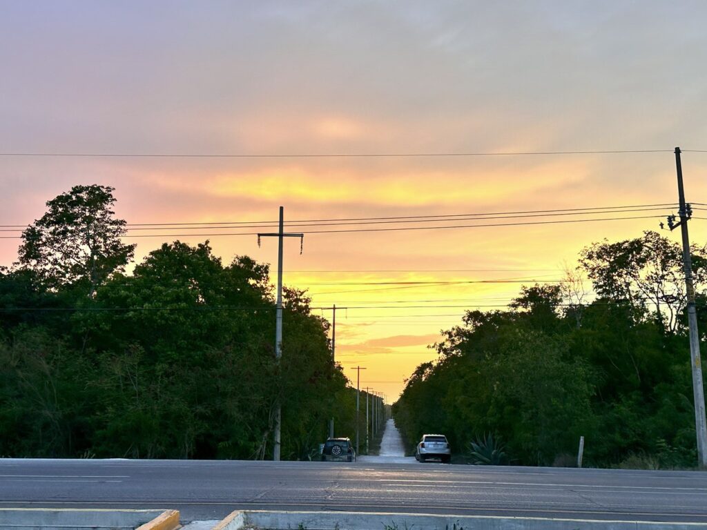 A view of land in Xpu Ha, Puerto Aventuras, with a sunset sky, trees along a narrow road, and parked cars, viewed from a roadside perspective.
