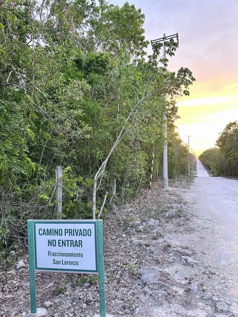 A dirt road in Xpu Ha, Puerto Aventuras, with a sign indicating a private, no-entry path to Fraccionamiento San Lorenzo, surrounded by dense green vegetation at sunset.