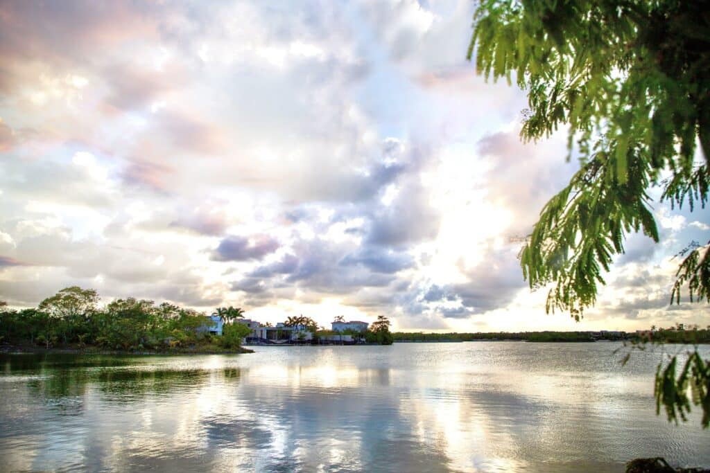 A scenic view of Lagos Del Sol 114 in Cancun, featuring a peaceful water body with lush greenery and modern houses along the Colosio Cancun area under a partly cloudy sky.