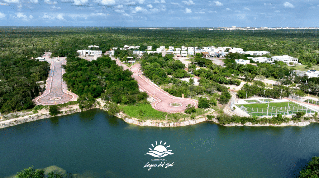 An aerial view of Lagos Del Sol 114 in Cancun's Colosio area, showing modern white houses, winding roads, a tennis court, and a lake, with lush green surroundings.