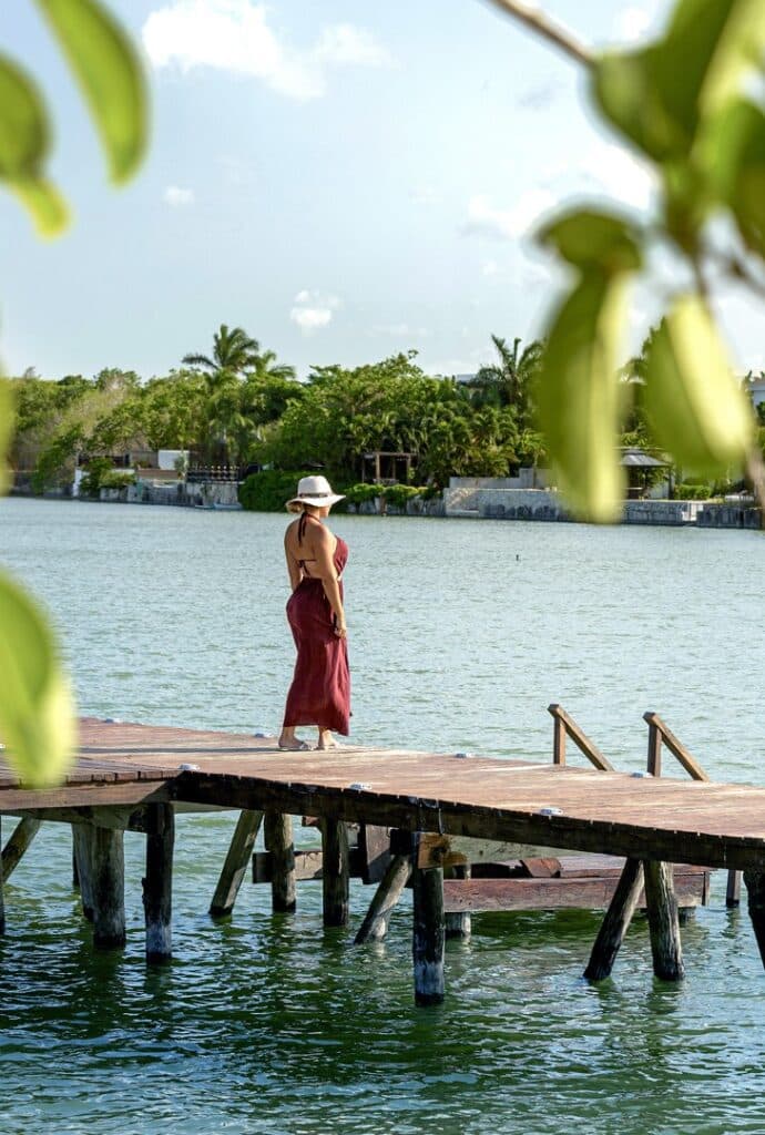 A woman in a red dress and wide-brimmed hat stands on a wooden dock overlooking a calm waterway in Colosio Cancun within Lagos Del Sol 114 development.