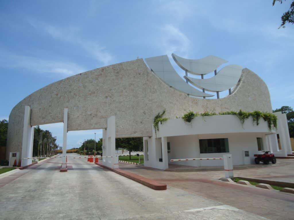 The entrance gate of Lagos Del Sol 114 in Cancun features a modern design with a curved stone and white structure topped with abstract white sculptures, set against a blue sky.