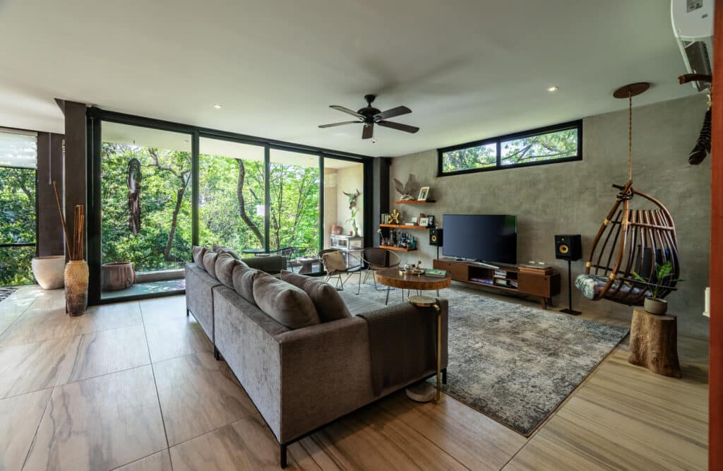 Living room in a multi-unit building at 3 Condos And Comercial space in Aldea Zama, Tulum, featuring large windows, a ceiling fan, a sectional sofa, a TV, and modern decor.