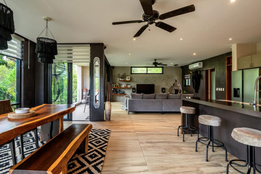 Interior view of a multi-unit building at 3 Condos and Commercial Space in Aldea Zama, Tulum, featuring a modern living area with a dining table, bar stools, and large windows opening to greenery.