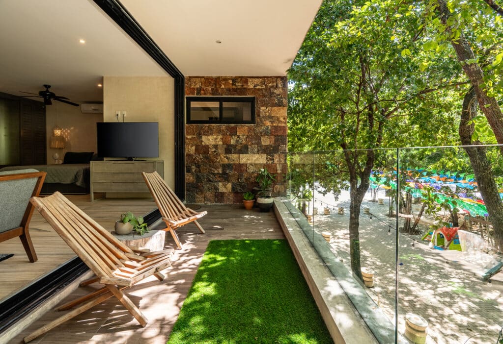 A spacious balcony with wooden lounge chairs and potted plants overlooks a lush green area with trees and colorful decorations in Aldea Zama, Tulum.