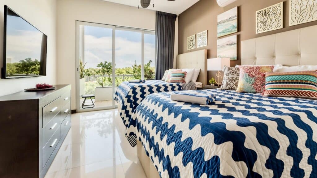 A bright bedroom in Oasis Penthouse, Playa del Carmen, with featuring blue and white patterned quilts and colorful pillows, a large glass door opening to a balcony with greenery view.