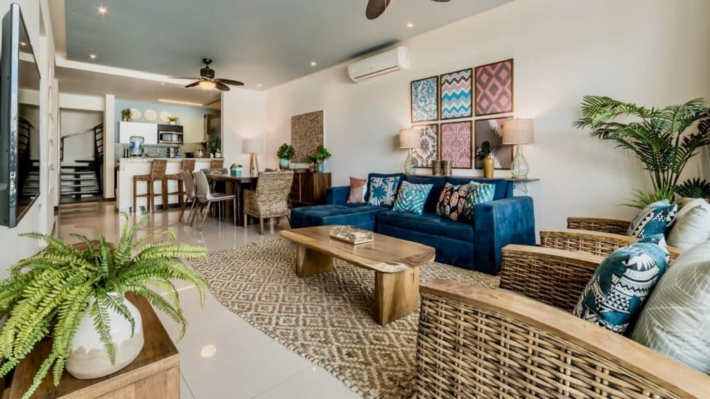Living room interior of Oasis Penthouse in Playa del Carmen, featuring a blue sofa, woven chairs, artwork, plants, and a dining area with a kitchen in the background.