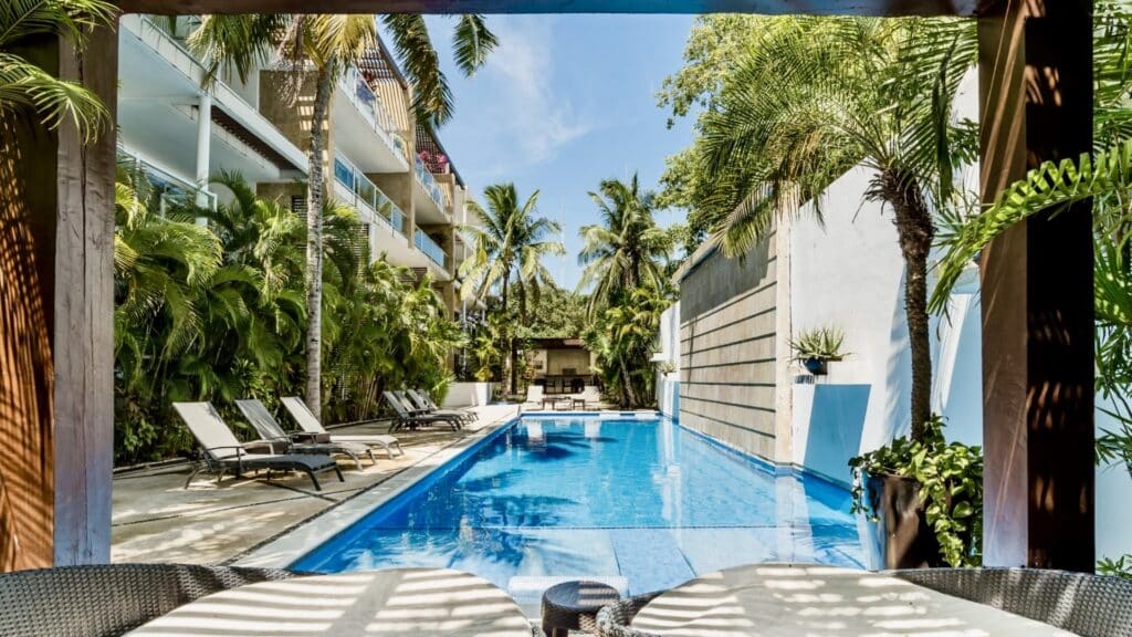 The Oasis Penthouse in Playa del Carmen features a pool surrounded by lush tropical plants and lounge chairs, with the city’s vibrant atmosphere just beyond.