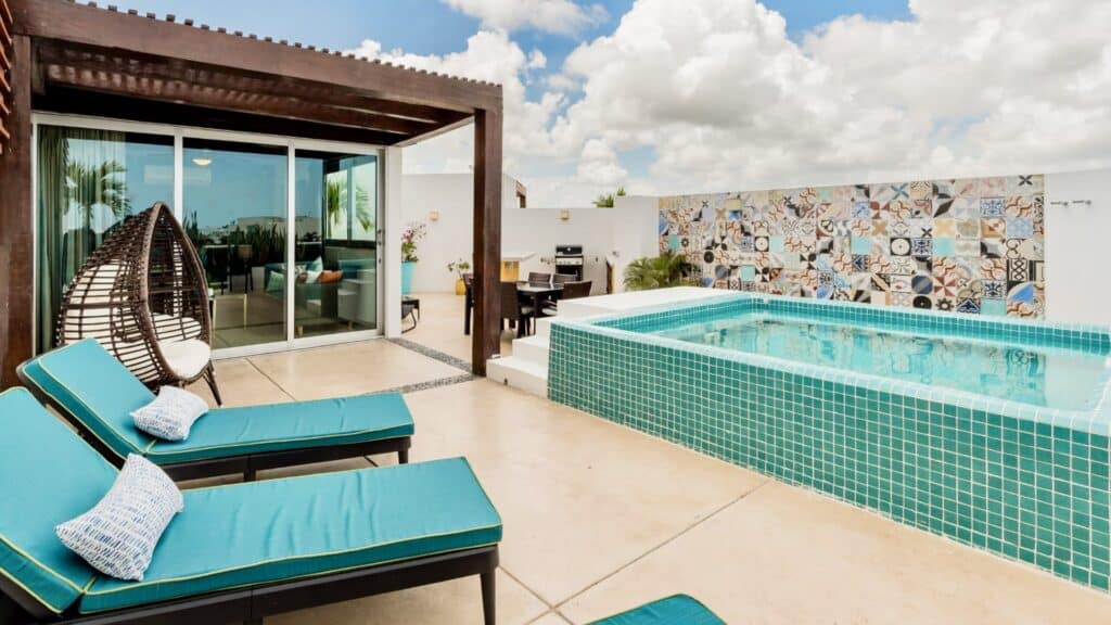 A rooftop terrace at Oasis Penthouse in Playa del Carmen features a small pool, two lounge chairs with teal cushions, a hanging wicker chair, and a colorful tiled wall under a partly cloudy sky.