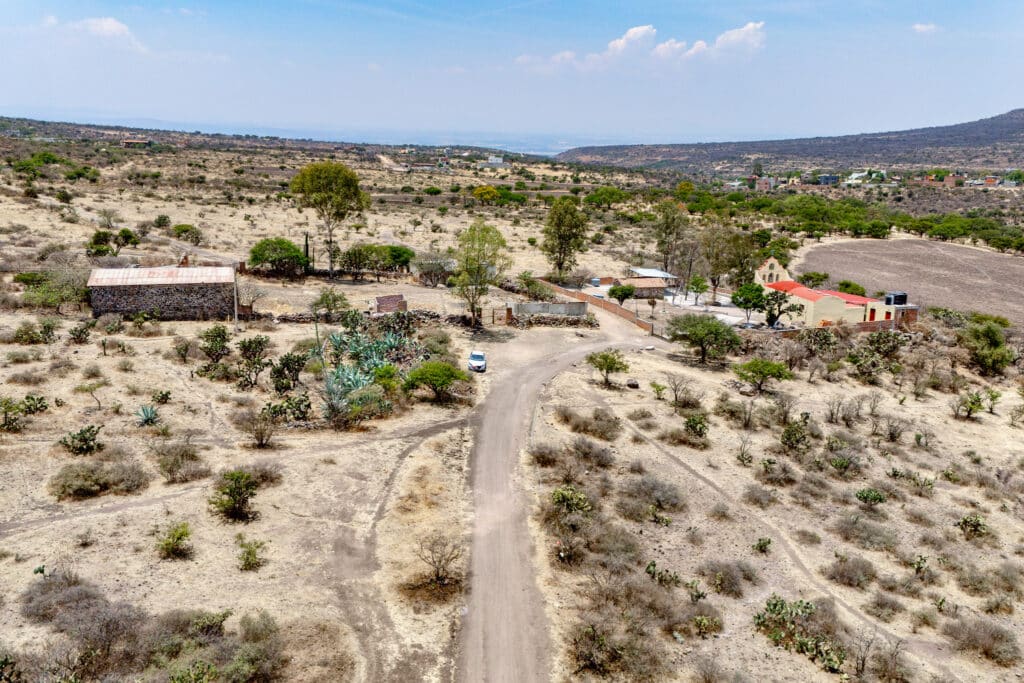 Aerial view of Rancho Guadalupe in San Miguel de Allende, showing a dry landscape with scattered trees, a small building, and a dirt driveway winding through the area.
