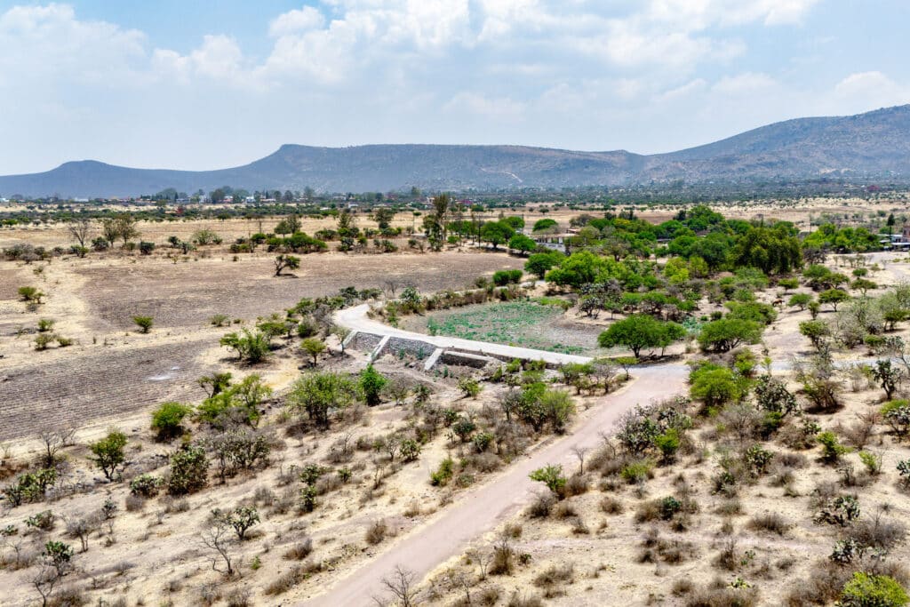Aerial view of Rancho Guadalupe in San Miguel de Allende, showing dry terrain with scattered trees, a dirt road, and distant mountains under a partly cloudy sky.