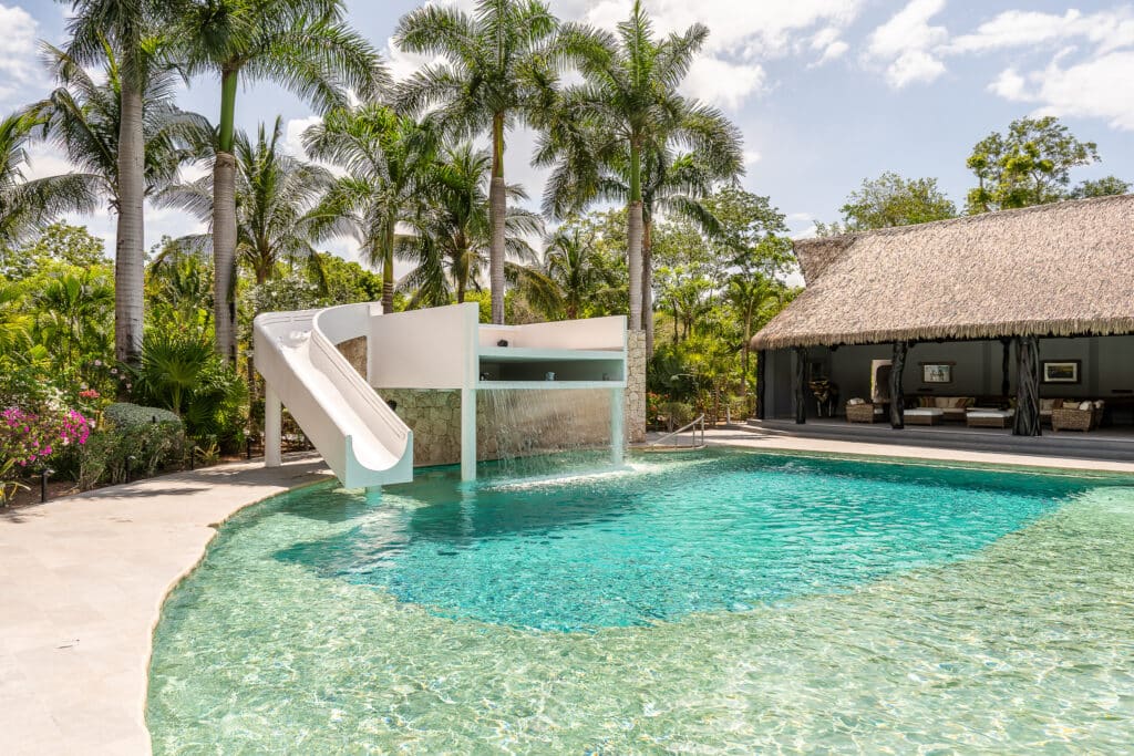 A swimming pool with a waterslide and waterfall feature is surrounded by lush tropical plants and tall palm trees, with a thatched-roof pavilion in the background in Akumal.