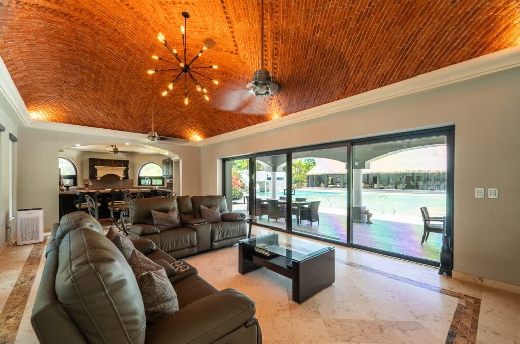Interior view of a spacious living room at Rancho Paraiso Oasis in Akumal, featuring leather sofas, a vaulted brick ceiling, and large sliding glass doors leading to a pool area.