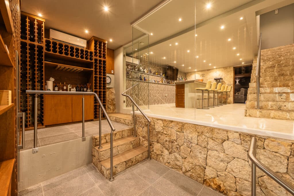 Interior view of Rancho Paraiso Oasis in Akumal, showing a stone-walled bar area with seating and a wine storage section, illuminated by recessed lighting.