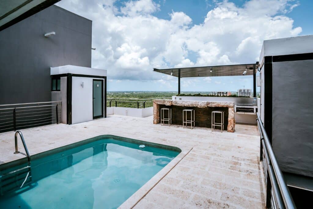 A rooftop view at Torre Onze Penthouse 3 in Merida, featuring a small swimming pool, bar area with stools, and an open sky with scattered clouds.