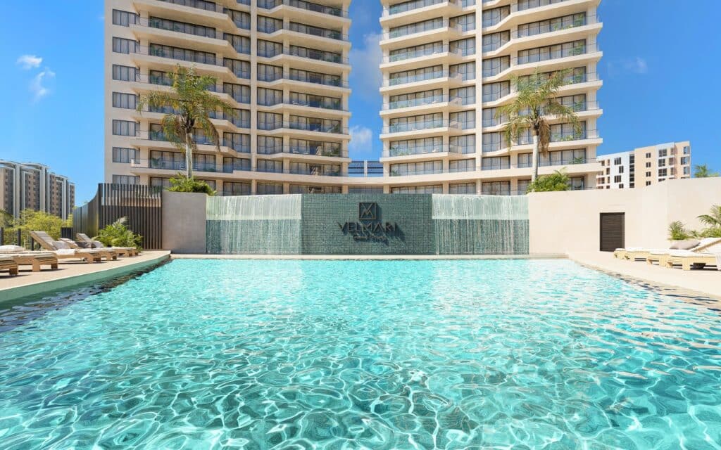 Swimming pool area in Velmari 604 development located in Puerto Cancun, with a modern high-rise building and surrounding sun loungers under a blue sky.