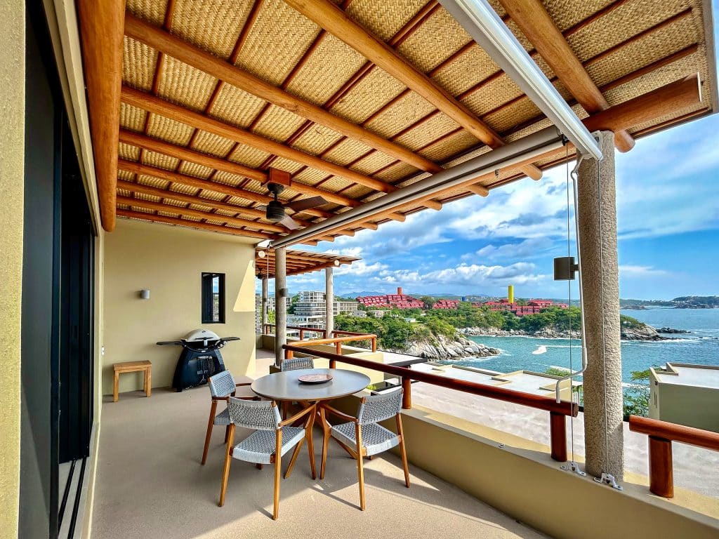 Outdoor balcony of Amanecer J2 in Playa Arrocito, Huatulco, featuring a round dining table, ceiling fan, grill, and overlooking the ocean with a scenic coastline and blue sky.