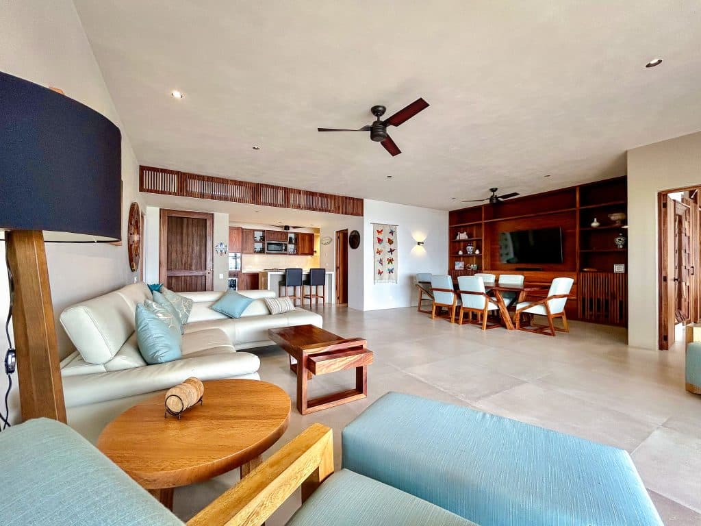 A spacious living area with a cream sofa and wooden furniture in the Amanecer J2 development near Playa Arrocito in Huatulco, featuring a large TV and open kitchen.
