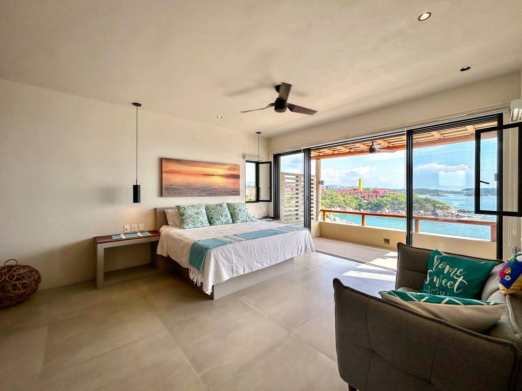 A bedroom in the Amanecer J2 development in Huatulco features a view of Playa Arrocito through large sliding glass doors, with a bed, sofa, and beach scenery outside.