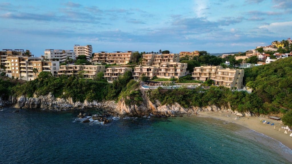 Amanecer J2 development in Playa Arrocito, Huatulco, features modern ocean-view penthouses perched on the rocky coastline with a small sandy beach and lush greenery surrounding the area.
