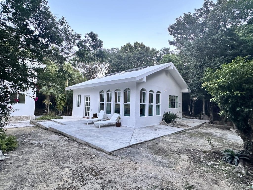 A white house with multiple arched and rectangular windows sits on Jungle Homes on 2.5 Acres in Puerto Aventuras, surrounded by lush trees and a simple outdoor patio area.