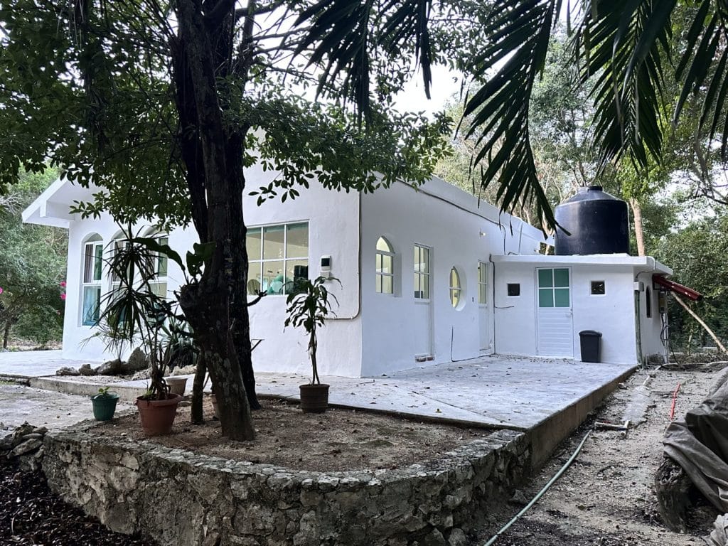 A white house with a modern design and multiple windows sits among lush trees and greenery in Jungle Homes on 2.5 Acres, Puerto Aventuras.