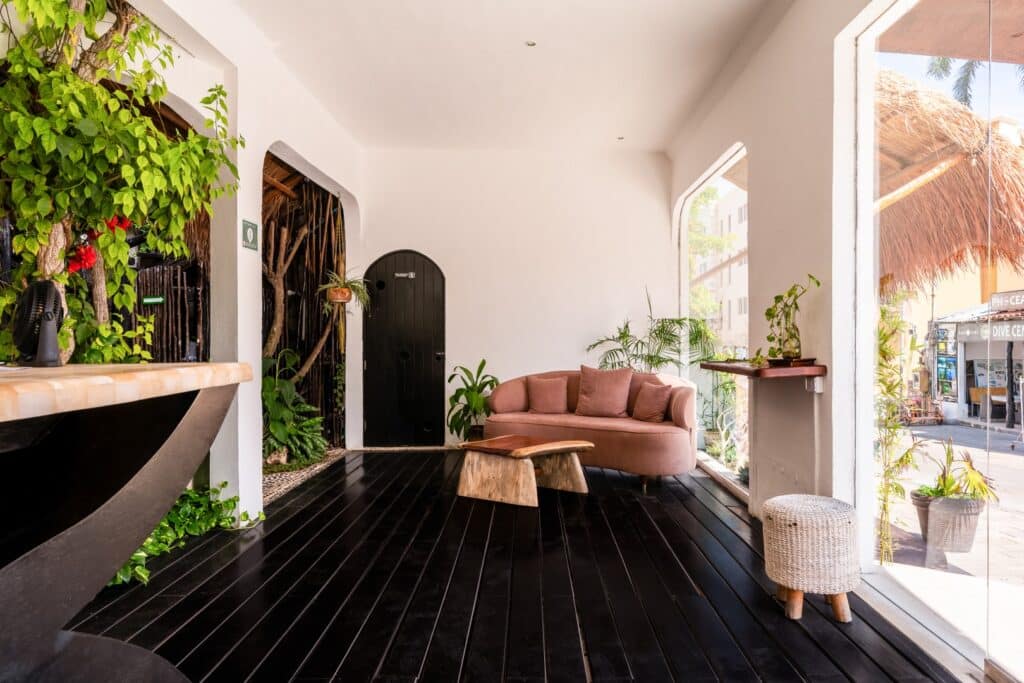 Interior of the Kinbe Hotel in Playa del Carmen's Centro, featuring a pink sofa with cushions, large windows with outdoor views, and decorative plants.