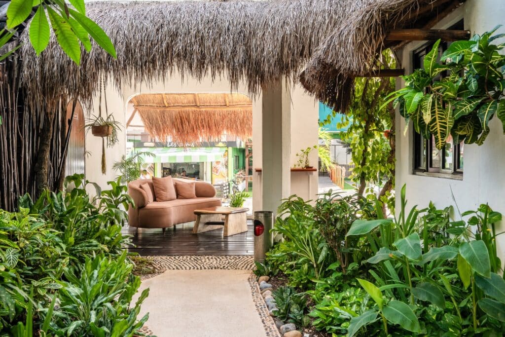 A cozy outdoor seating area at the Kinbe Hotel in Playa del Carmen's Centro, featuring lush greenery, a thatched roof, and a comfortable pink sofa.