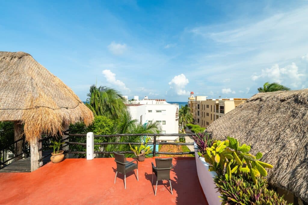 A view from the terrace of the Kinbe Hotel in Playa del Carmen's Centro area, featuring two thatched roofs, colorful plants, and a glimpse of the cityscape and ocean in the background.